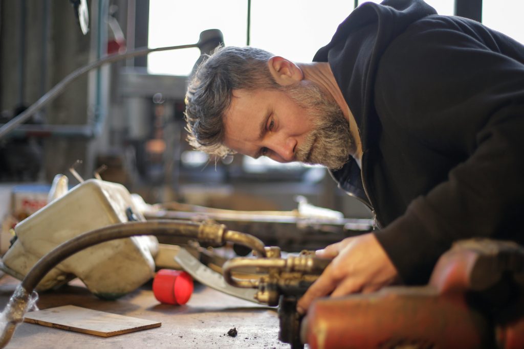 Side view of serious brutal mechanic in workwear standing at workbench and fixing metal detail while working in garage